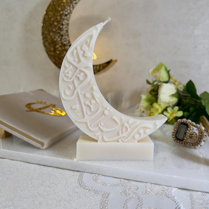 Künstlerisch gestaltete Ramadan-Halbmondkerze aus weißem Wachs mit detaillierter arabischer Kalligrafie. Die Kerze steht auf einer stabilen Basis und wird von eleganten goldfarbenen Dekorationselementen, Blumen und textilen Unterlagen ergänzt. Die harmonische Farbkomposition und das sanfte Licht unterstreichen den festlichen Charakter und machen diese Kerze ideal für spirituelle Dekorationen im Ramadan.