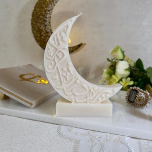 Detailreiche Ramadan-Halbmondkerze in Weiß mit kunstvoll eingearbeiteter arabischer Schrift. Die Kerze ist freistehend auf einer rechteckigen Basis platziert und von goldenen Akzenten, Blumenarrangements und feinen Stofftexturen umgeben. Die Kombination aus minimalistischer Eleganz und spiritueller Symbolik macht diese Kerze zu einem besonderen Blickfang für festliche Tischdekorationen während des Ramadans.