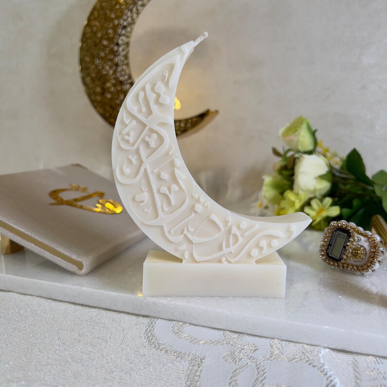 Detailreiche Ramadan-Halbmondkerze in Weiß mit kunstvoll eingearbeiteter arabischer Schrift. Die Kerze ist freistehend auf einer rechteckigen Basis platziert und von goldenen Akzenten, Blumenarrangements und feinen Stofftexturen umgeben. Die Kombination aus minimalistischer Eleganz und spiritueller Symbolik macht diese Kerze zu einem besonderen Blickfang für festliche Tischdekorationen während des Ramadans.