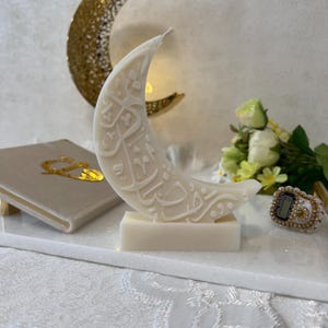 Elegante Halbmondkerze aus weißem Wachs mit aufwendig gestalteter arabischer Kalligrafie, dekorativ in einer festlichen Ramadan-Szene arrangiert. Goldene Monddekorationen, florale Elemente und sanfte Lichtreflexionen schaffen eine warme, spirituelle Stimmung. Diese Kerze verbindet moderne Ästhetik mit traditioneller Bedeutung und eignet sich ideal als stilvolle Ramadan-Dekoration oder Geschenkidee.