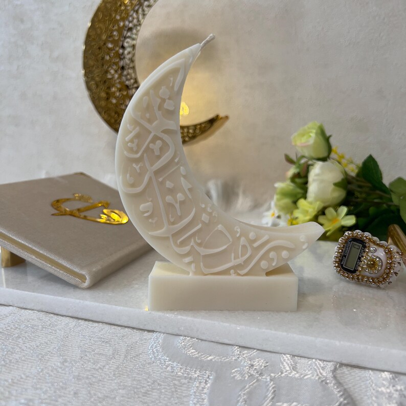 Elegante Halbmondkerze aus weißem Wachs mit aufwendig gestalteter arabischer Kalligrafie, dekorativ in einer festlichen Ramadan-Szene arrangiert. Goldene Monddekorationen, florale Elemente und sanfte Lichtreflexionen schaffen eine warme, spirituelle Stimmung. Diese Kerze verbindet moderne Ästhetik mit traditioneller Bedeutung und eignet sich ideal als stilvolle Ramadan-Dekoration oder Geschenkidee.