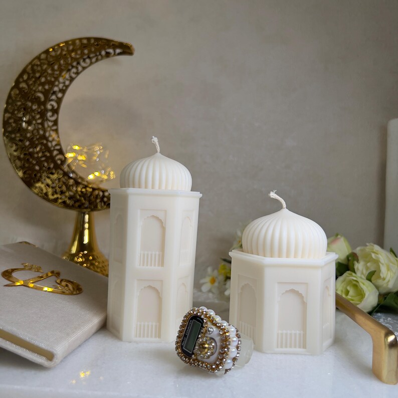 Ramadan Deko Kerzen Set mit zwei cremeweißen Moschee-Kerzen aus Wachs, eine hohe Turmkerze und eine niedrigere sechseckige Kerze mit geriffelter Kuppel. Orientalische Fensterdetails, eleganter Halbmond-Dekor im Hintergrund, Blumen und Gebetsaccessoires. Ideal als islamische Wohndeko, Ramadan Tischdeko oder stilvolle Eid Geschenkidee.