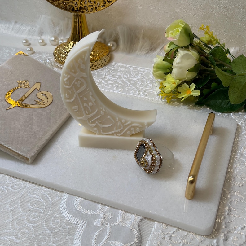 Minimalistische Ramadan-Dekokerze in Halbmondform mit kunstvoller arabischer Kalligrafie. Die weiße Kerze ruht auf einem schlichten Sockel und wird von goldenen Dekorationselementen, Blumen und weichen Textilien stilvoll ergänzt. Das ruhige, helle Farbkonzept vermittelt Spiritualität und Eleganz und eignet sich perfekt als festliche Dekoration während des Fastenmonats Ramadan.