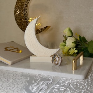 Edle weiße Halbmond-Kerze mit arabischer Kalligrafie, dekorativ arrangiert auf einer hellen Oberfläche. Im Hintergrund sorgen goldene Mondornamente, zarte Blumen und warme Lichtakzente für eine festliche Ramadan-Stimmung. Die klaren Linien der Kerze und die feinen Schriftdetails verbinden modernes Design mit traditioneller Symbolik und schaffen eine ruhige, spirituelle Atmosphäre für besondere Anlässe.