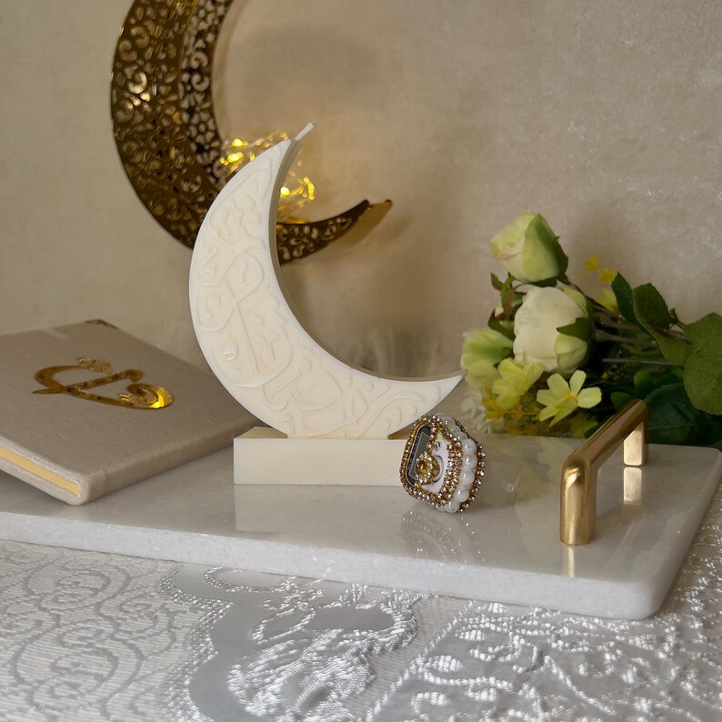 Edle weiße Halbmond-Kerze mit arabischer Kalligrafie, dekorativ arrangiert auf einer hellen Oberfläche. Im Hintergrund sorgen goldene Mondornamente, zarte Blumen und warme Lichtakzente für eine festliche Ramadan-Stimmung. Die klaren Linien der Kerze und die feinen Schriftdetails verbinden modernes Design mit traditioneller Symbolik und schaffen eine ruhige, spirituelle Atmosphäre für besondere Anlässe.