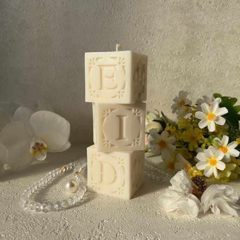 May include: Three ivory-coloured, square candles stacked to spell "EID". Each candle has a letter and floral design. The candles are on a white surface, with flowers and a pearl necklace in the background.