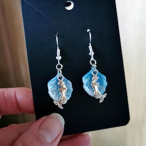 Peut inclure: Une paire de boucles d'oreilles sirène. Chaque boucle d'oreille présente un charm sirène argenté suspendu à un crochet argenté. La sirène est fixée à une base bleue clair en forme de coquillage. Les boucles d'oreilles sont présentées sur une carte noire.