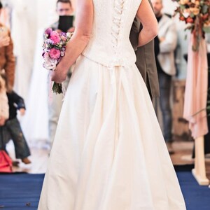 Puede incluir: Una novia con un vestido de novia blanco con un corpiño de encaje y una falda amplia. La parte trasera del vestido presenta un cierre tipo corsé. Sostiene un ramo de flores rosas y moradas. La ceremonia se lleva a cabo en interiores.