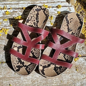 May include: A pair of open-toe sandals with a snakeskin pattern footbed and burgundy leather straps. The sandals are displayed on a weathered wooden surface, with yellow flower petals scattered around.
