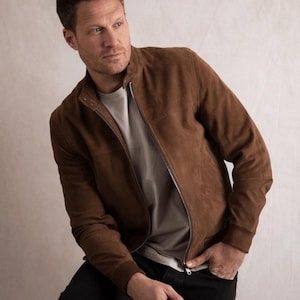 May include: A brown suede bomber jacket with a zipper closure. The jacket has a classic design and a collarless neckline. The model is wearing a gray t-shirt and black jeans. The jacket is a medium brown color.