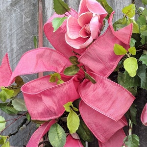 May include: A pink magnolia wreath with a large matching bow. The wreath features pink magnolia flowers, green leaves, and a woven grapevine base. The bow's ribbon cascades down the wreath, adding a touch of elegance.
