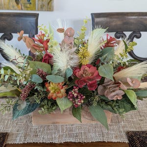 May include: A rectangular wooden planter box filled with an arrangement of artificial greenery, including red and green succulents, feathery cream-colored pampas grass plumes, eucalyptus leaves, and small clusters of red berries.