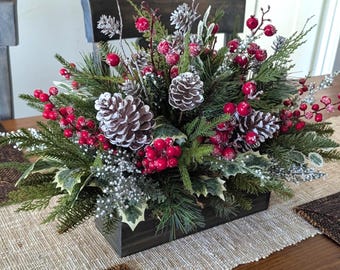 May include: A rectangular dark wooden box filled with artificial greenery, including pine branches and holly leaves. The arrangement features clusters of bright red berries and several pinecones with white-tipped edges, creating a winter-themed display.