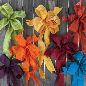 May include: Seven different colored silk ribbon bows hanging on a wooden fence. The bows are in shades of green, orange, yellow, red, purple, and blue.