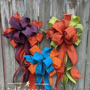 May include: Three large, decorative bows made from two different colored ribbons. The bows are tied together and hang from a wooden fence. The ribbons are orange, teal, purple, and green. The text "TWO RIBBON BOW" is written in yellow on the fence.