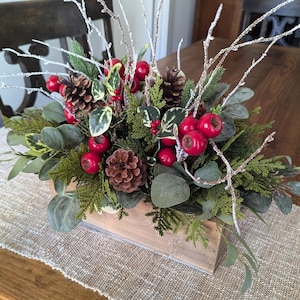 May include: A wooden box filled with a festive Christmas centerpiece. The arrangement features red berries, pine cones, evergreen branches, and frosted twigs.
