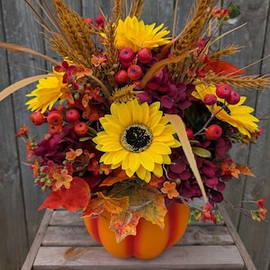 May include: A fall-themed floral arrangement in an orange pumpkin-shaped vase. The arrangement features bright yellow sunflowers, red berries, and burgundy flowers, with wheat stalks and fall leaves. The vase sits on a wooden surface.