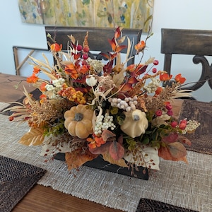 May include: A floral centerpiece in a dark rectangular wooden box featuring two small artificial pumpkins, orange and red autumn leaves, dried grasses, white berry clusters, and small red berries arranged in a dense, multi-textured display.
