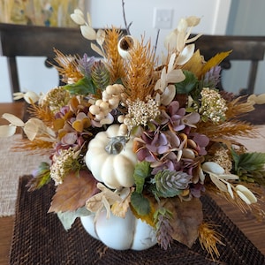 May include: A fall-themed floral arrangement in a white pumpkin-shaped vase. The arrangement includes dried wheat, purple hydrangeas, and various fall foliage. The vase sits on a brown placemat on a wooden table.