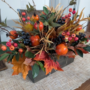 May include: A fall-themed floral arrangement in a rectangular wooden box. The arrangement features orange and red berries, orange-colored fruit, and colorful fall leaves. The arrangement is on a beige tablecloth.