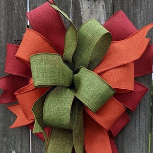 May include: A large, decorative bow made with three different colored ribbons. The bow is made with a green, orange, and burgundy ribbon. The bow is tied in a knot and has a long tail.