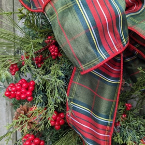 May include: A green and red plaid ribbon with a red trim is tied into a bow on a Christmas wreath. The wreath is decorated with pine branches, red berries, and pine cones.