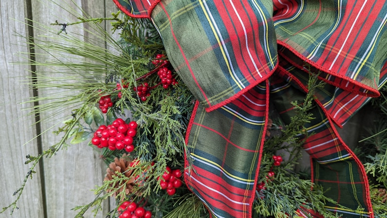May include: A green and red plaid ribbon with a red trim is tied into a bow on a Christmas wreath. The wreath is decorated with pine branches, red berries, and pine cones.