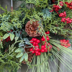 May include: A Christmas wreath with green pine branches, red berries, and a brown pine cone. The wreath is decorated with a plaid ribbon.
