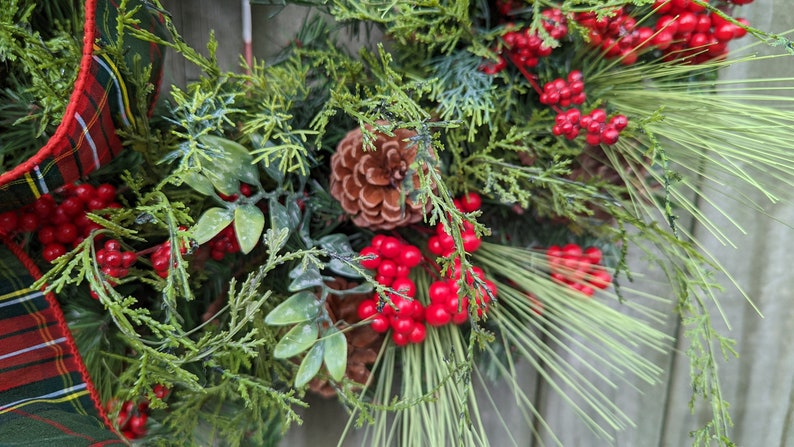 May include: A Christmas wreath with green pine branches, red berries, and a brown pine cone. The wreath is decorated with a plaid ribbon.