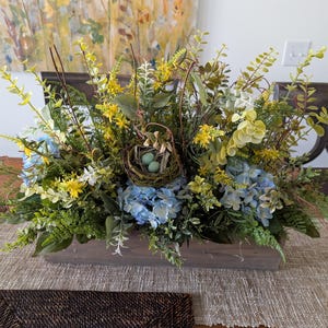 May include: A wooden box centerpiece filled with a variety of greenery, yellow and blue flowers, and a bird's nest with blue eggs.