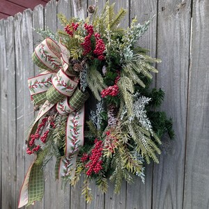 May include: A festive Christmas wreath with a large bow, red berries, and pinecones. The bow features a green and cream plaid pattern with red berry accents. The wreath is made of evergreen branches and is displayed on a weathered wooden fence.