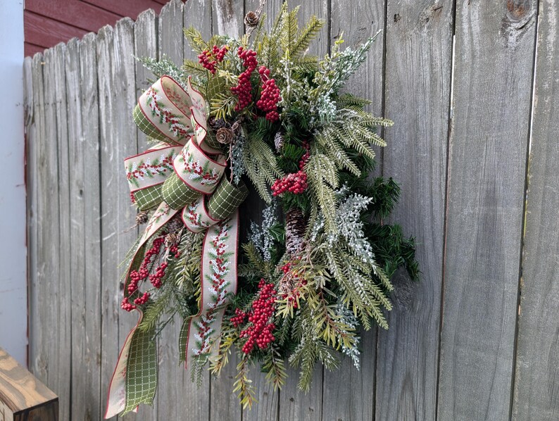 May include: A festive Christmas wreath with a large bow, red berries, and pinecones. The bow features a green and cream plaid pattern with red berry accents. The wreath is made of evergreen branches and is displayed on a weathered wooden fence.