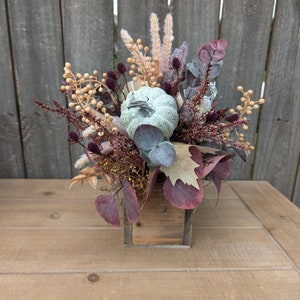 May include: A fall-themed floral arrangement in a small, square wooden box. The arrangement features a light green pumpkin, burgundy leaves, and various dried flowers and berries in shades of beige, brown, and purple. The background is a wooden fence.