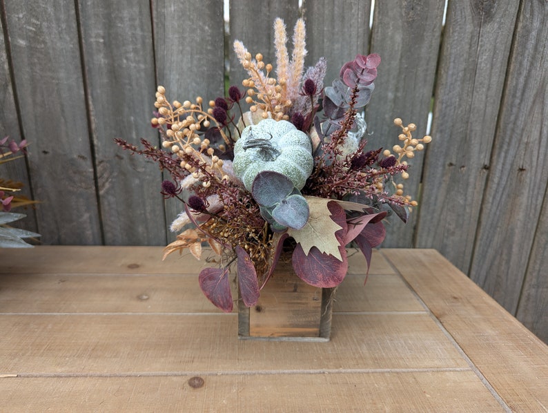 May include: A fall-themed floral arrangement in a small, square wooden box. The arrangement features a light green pumpkin, burgundy leaves, and various dried flowers and berries in shades of beige, brown, and purple. The background is a wooden fence.