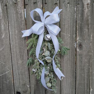 May include: A decorative teardrop-shaped swag featuring frosted green pine needles and eucalyptus leaves. The arrangement is accented with small silver jingle bells and topped with a large, textured white ribbon tied in a bow.