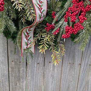 May include: A festive Christmas wreath with vibrant red berries and lush green pine needles. A decorative ribbon with a berry and pine needle pattern adds a touch of elegance. The wreath is set against a rustic wooden background.