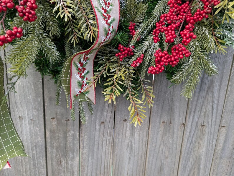 May include: A festive Christmas wreath with vibrant red berries and lush green pine needles. A decorative ribbon with a berry and pine needle pattern adds a touch of elegance. The wreath is set against a rustic wooden background.