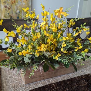 May include: A wooden box planter filled with a lush arrangement of yellow flowers and green foliage. The planter is sitting on a table with a brown placemat.