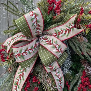 May include: A festive Christmas wreath featuring a large bow with a beige ribbon adorned with red berries and green sprigs. The bow has a green diamond pattern on the edges. The wreath is decorated with red berries, pinecones, and evergreen branches.