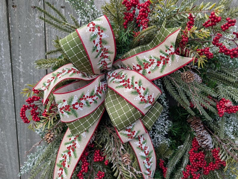 May include: A festive Christmas wreath featuring a large bow with a beige ribbon adorned with red berries and green sprigs. The bow has a green diamond pattern on the edges. The wreath is decorated with red berries, pinecones, and evergreen branches.
