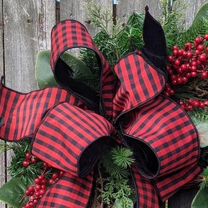 May include: A festive wreath featuring a large bow made of red and black buffalo plaid ribbon. The wreath is adorned with green foliage, red berries, and magnolia leaves, creating a rustic holiday decoration.