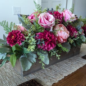 May include: A wooden box filled with a floral arrangement of pink and burgundy flowers. The arrangement includes roses, peonies, and dahlias.