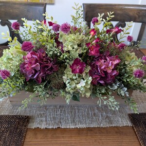 Pu&ograve; includere: Centrotavola rettangolare in legno con fiori artificiali. Include ortensie viola scuro, cardi rosa e fogliame verde e bianco. Il centrotavola &egrave; posizionato su un runner da tavolo beige.