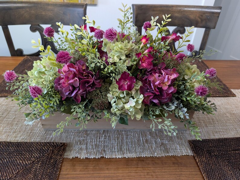 Pu&ograve; includere: Centrotavola rettangolare in legno con fiori artificiali. Include ortensie viola scuro, cardi rosa e fogliame verde e bianco. Il centrotavola &egrave; posizionato su un runner da tavolo beige.