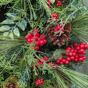 May include: Close-up of a Christmas wreath with green pine needles, red berries, and a brown pine cone.