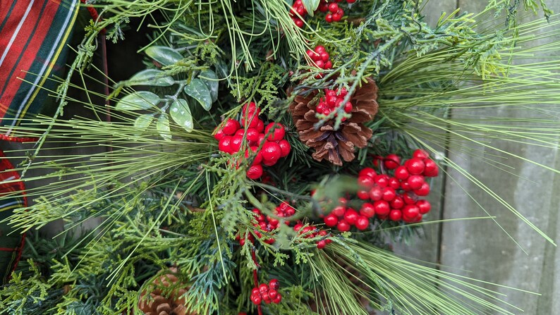 May include: Close-up of a Christmas wreath with green pine needles, red berries, and a brown pine cone.