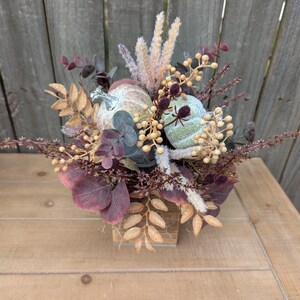 May include: A fall-themed floral arrangement in a small wooden container. The arrangement features two small, light green pumpkins, burgundy eucalyptus leaves, and beige and gold accents. The arrangement sits on a light brown wooden table.