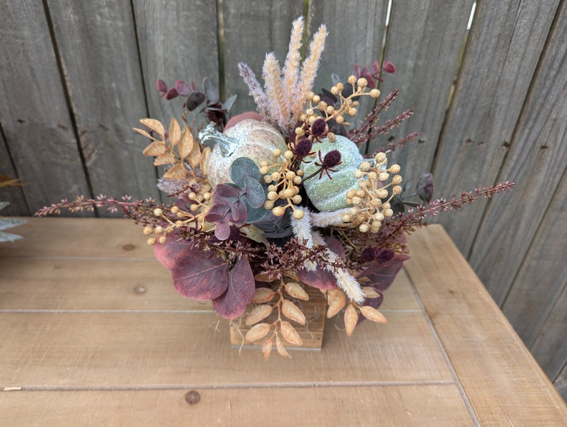 May include: A fall-themed floral arrangement in a small wooden container. The arrangement features two small, light green pumpkins, burgundy eucalyptus leaves, and beige and gold accents. The arrangement sits on a light brown wooden table.