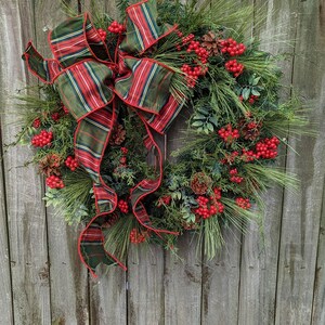 May include: A Christmas wreath with a plaid bow, pine branches, red berries, and pine cones. The wreath is hanging on a wooden fence.