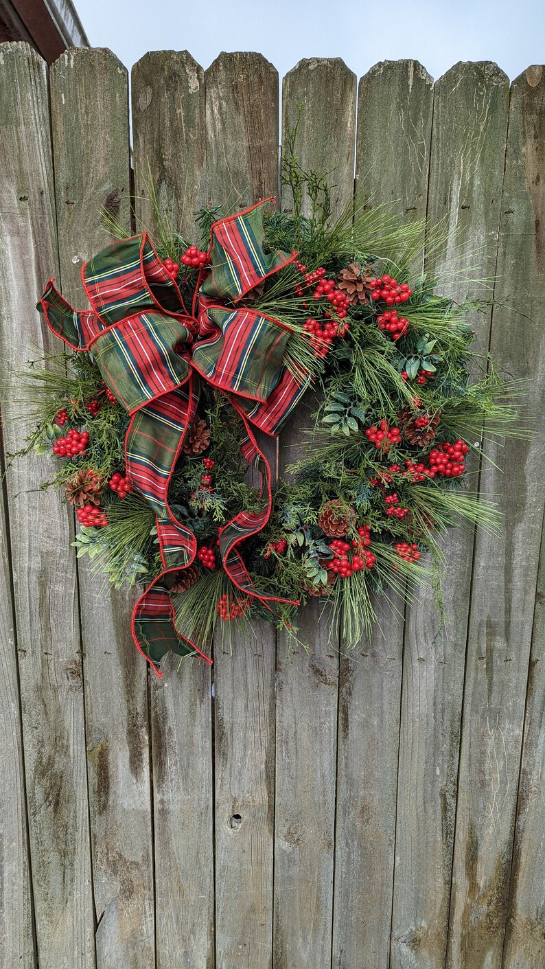 May include: A Christmas wreath with a plaid bow, pine branches, red berries, and pine cones. The wreath is hanging on a wooden fence.